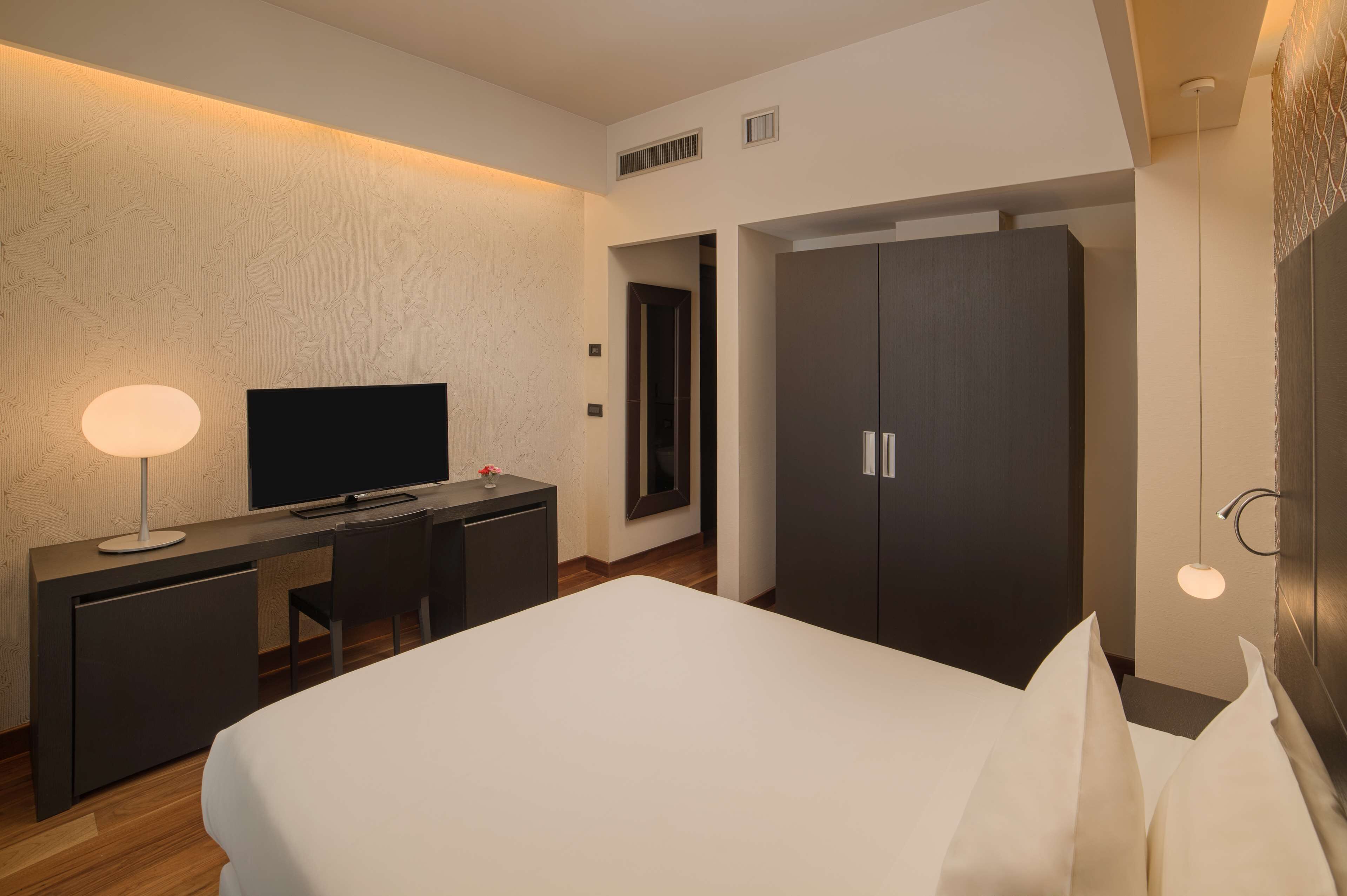 A hotel room with a bed, a desk with a TV, a wardrobe, and a lamp.