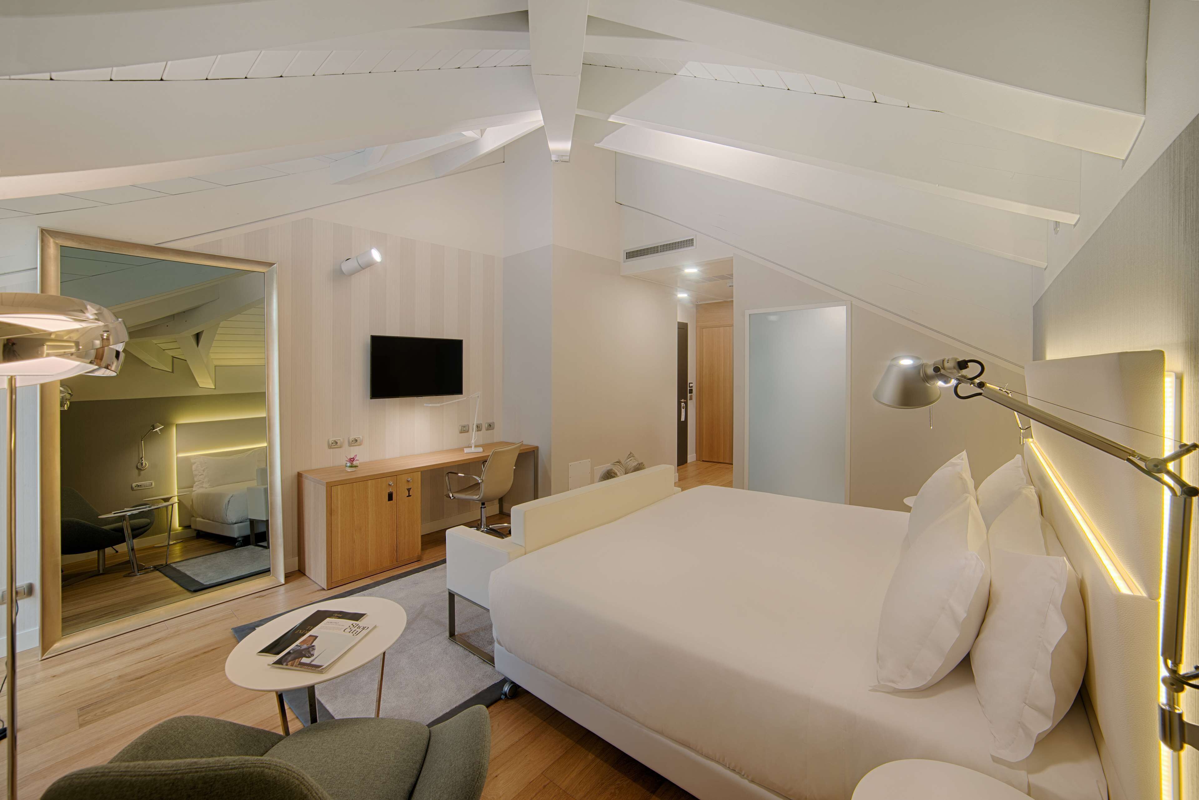 Ein modernes Hotelzimmer mit einem großen Bett, einem Schreibtisch, einem Sessel, einem Fernseher und einem begehbaren Kleiderschrank.