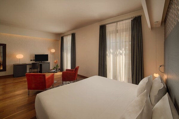 A hotel room with a large bed, a desk with a TV, two red chairs, and a large window with curtains.