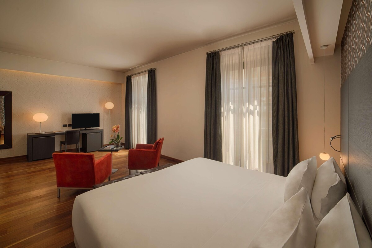 A hotel room with a large bed, a desk with a TV, two red chairs, and a desk lamp.