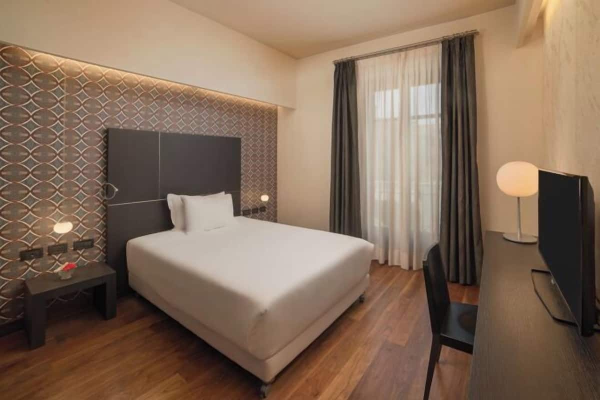 Ein Hotelzimmer mit einem großen Bett, einem Schreibtisch mit Stuhl, einem Fernseher und einem Fenster mit Vorhängen.