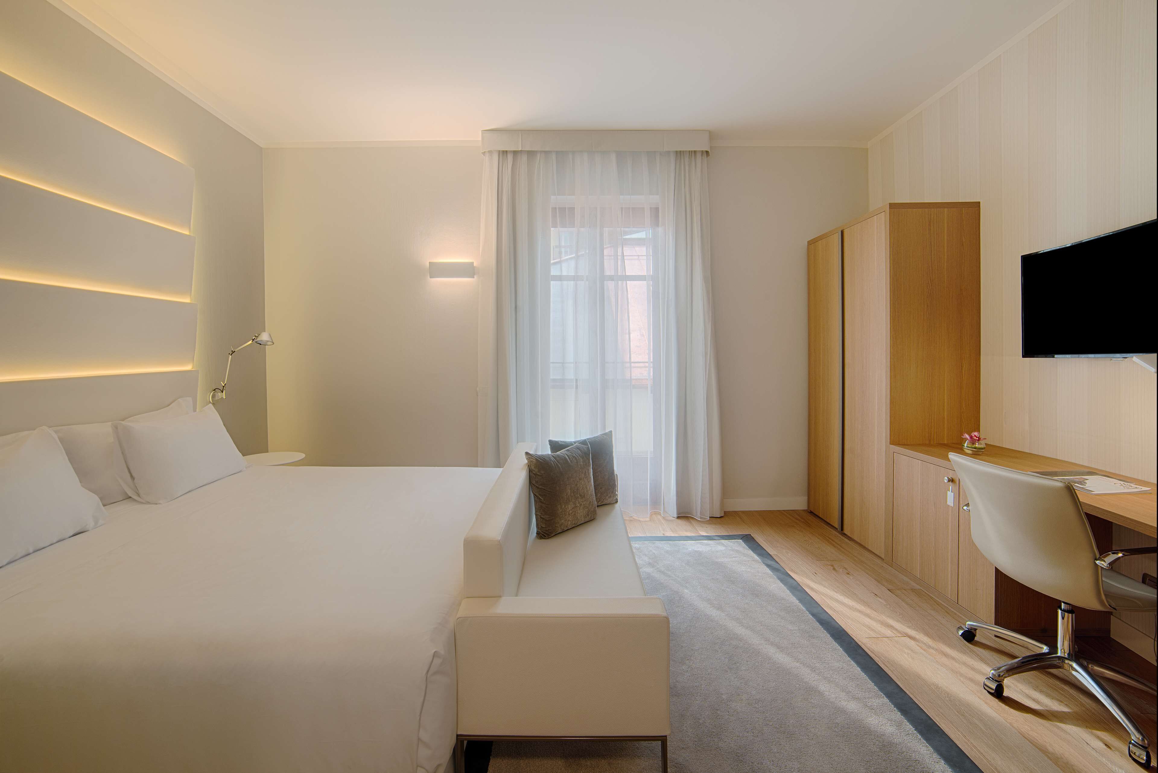 A modern hotel room with a large bed, a sitting area, a desk, and a TV.