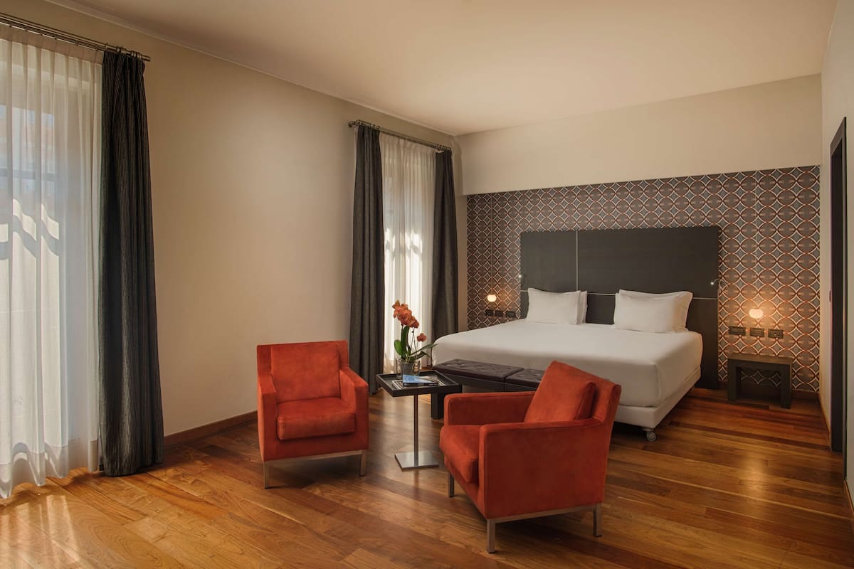 Ein Hotelzimmer mit einem großen Bett, zwei roten Sessel, einem kleinen Tisch mit Blumenvase und einem Nachttisch mit Lampe.