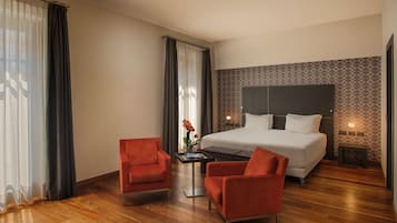 Quarto de hotel com uma cama grande, duas poltronas vermelhas, uma mesa pequena com um vaso de flores e uma mesa de cabeceira com abajur.
