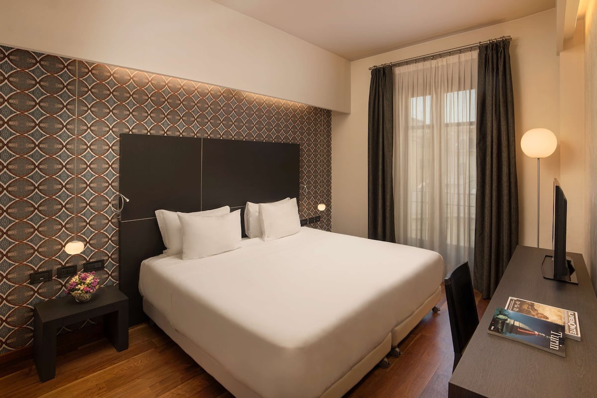 Ein modernes Hotelzimmer mit einem großen Bett, einem Nachttisch mit Lampe, einem kleinen Tisch mit Blumenvase und einer Zeitschrift auf dem Beistelltisch.