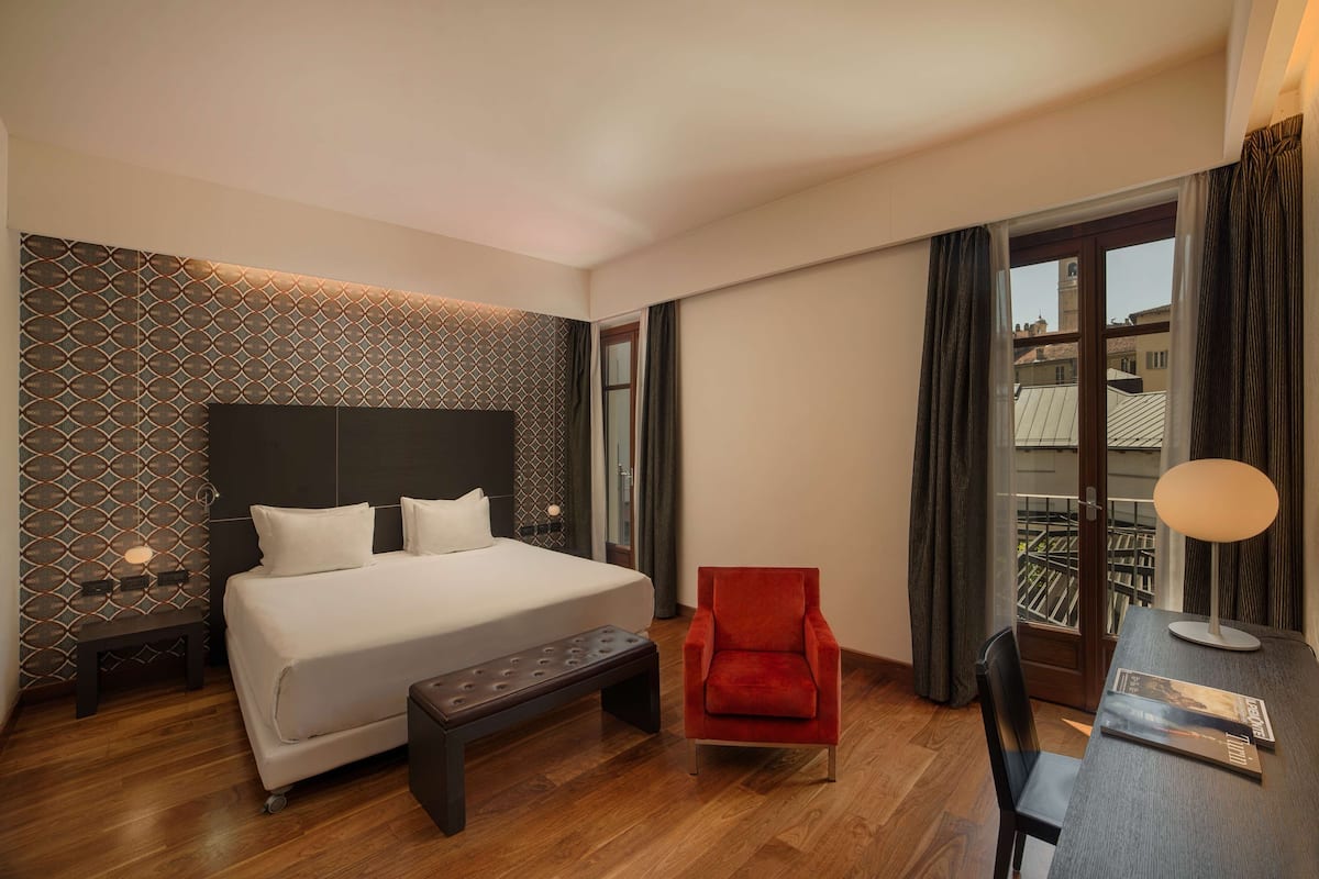 Ein Hotelzimmer mit einem großen Bett, einem roten Sessel, einem Schreibtisch mit Lampe und einer Wand mit Muster.