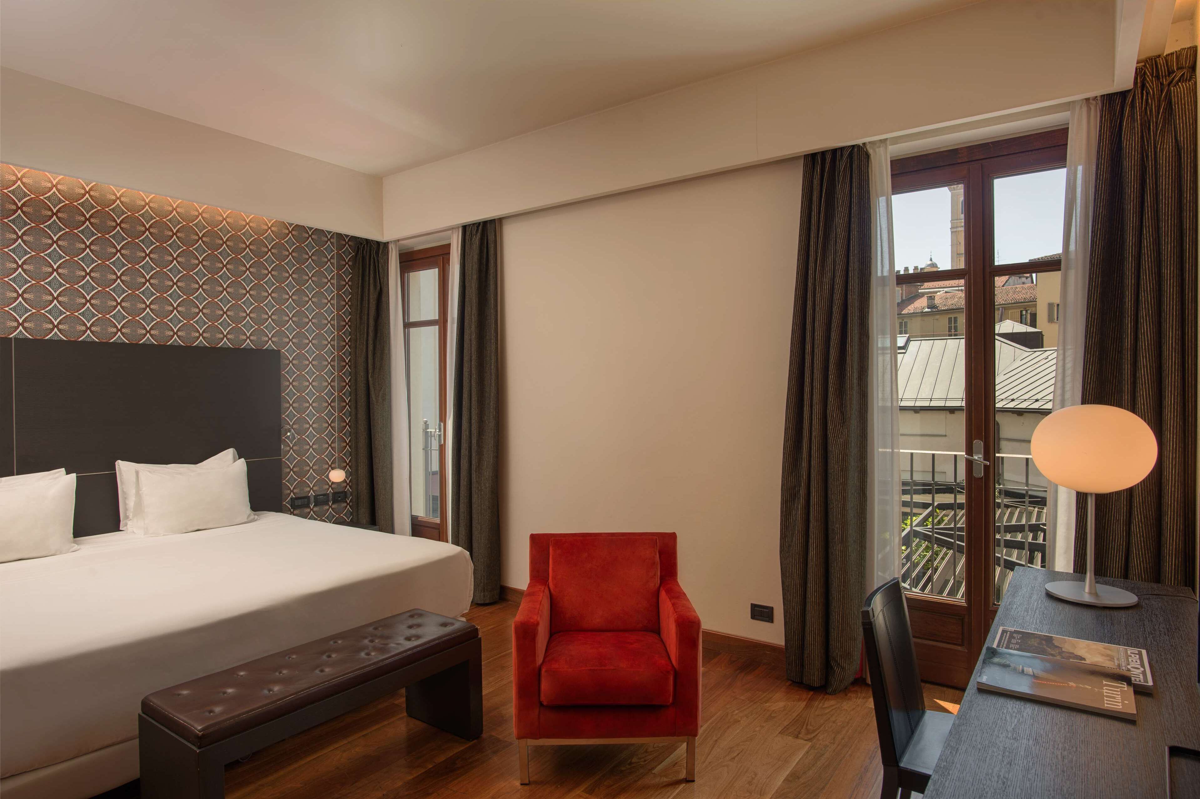 Un cuarto de hotel con una cama grande, un sillón rojo, un banco de madera, un escritorio con una lámpara y vistas a edificios a través de una ventana.