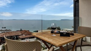 Une table en bois, un verre de vin rouge et un sac noir sont posés sur un balcon donnant sur une étendue d’eau.