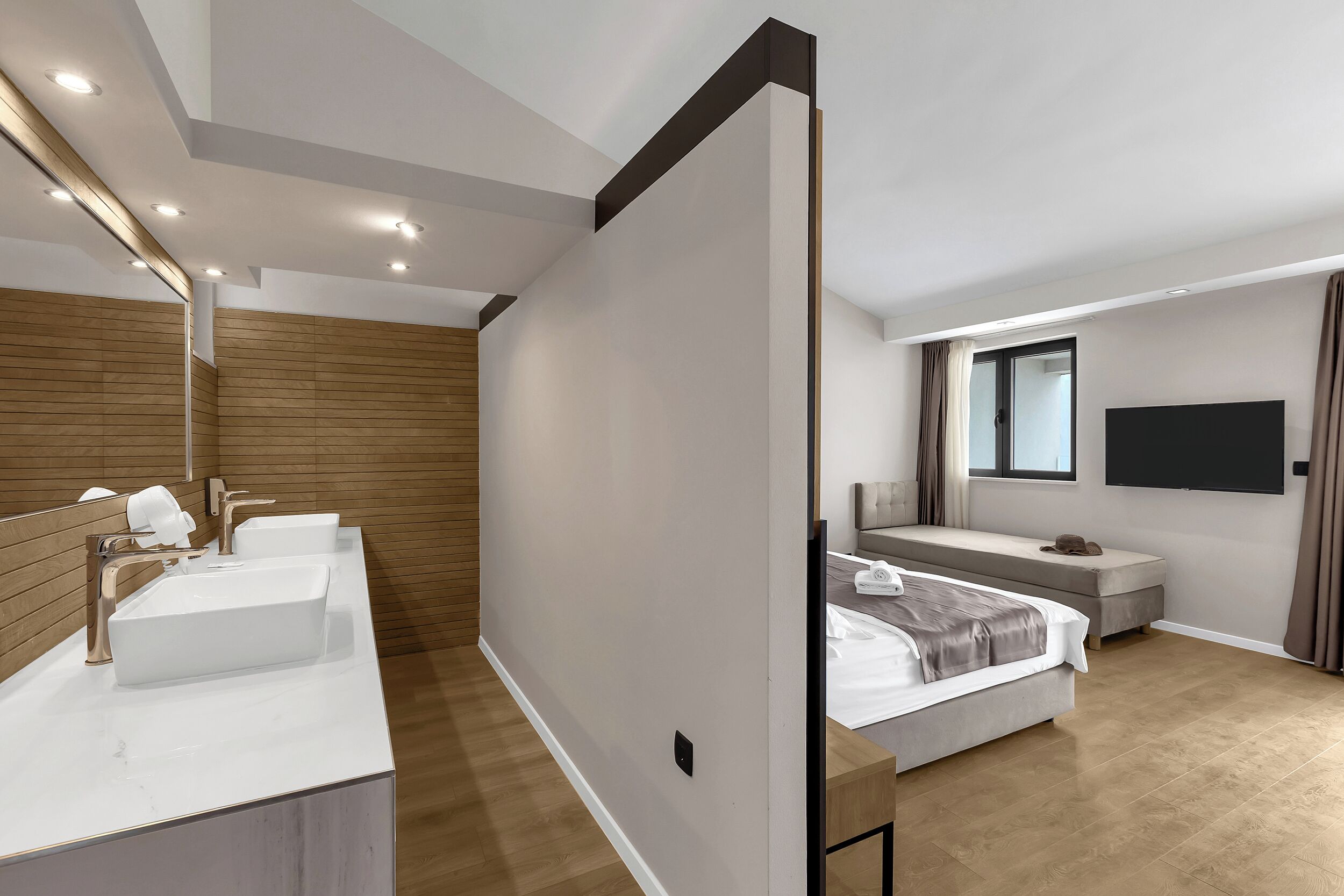 A modern hotel room with a large bed, a TV mounted on the wall, and a separate bathroom with a vanity and two sinks.