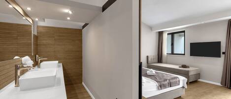 A modern hotel room with a large bed, a TV mounted on the wall, and a separate bathroom with a vanity and two sinks.