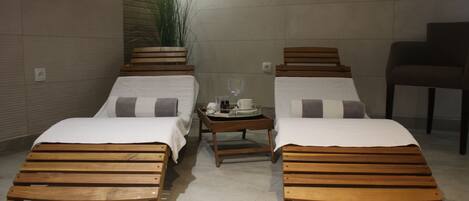 Duas camas de madeira com roupa de cama branca, uma mesa pequena com um tabuleiro de objetos e uma planta em vaso.