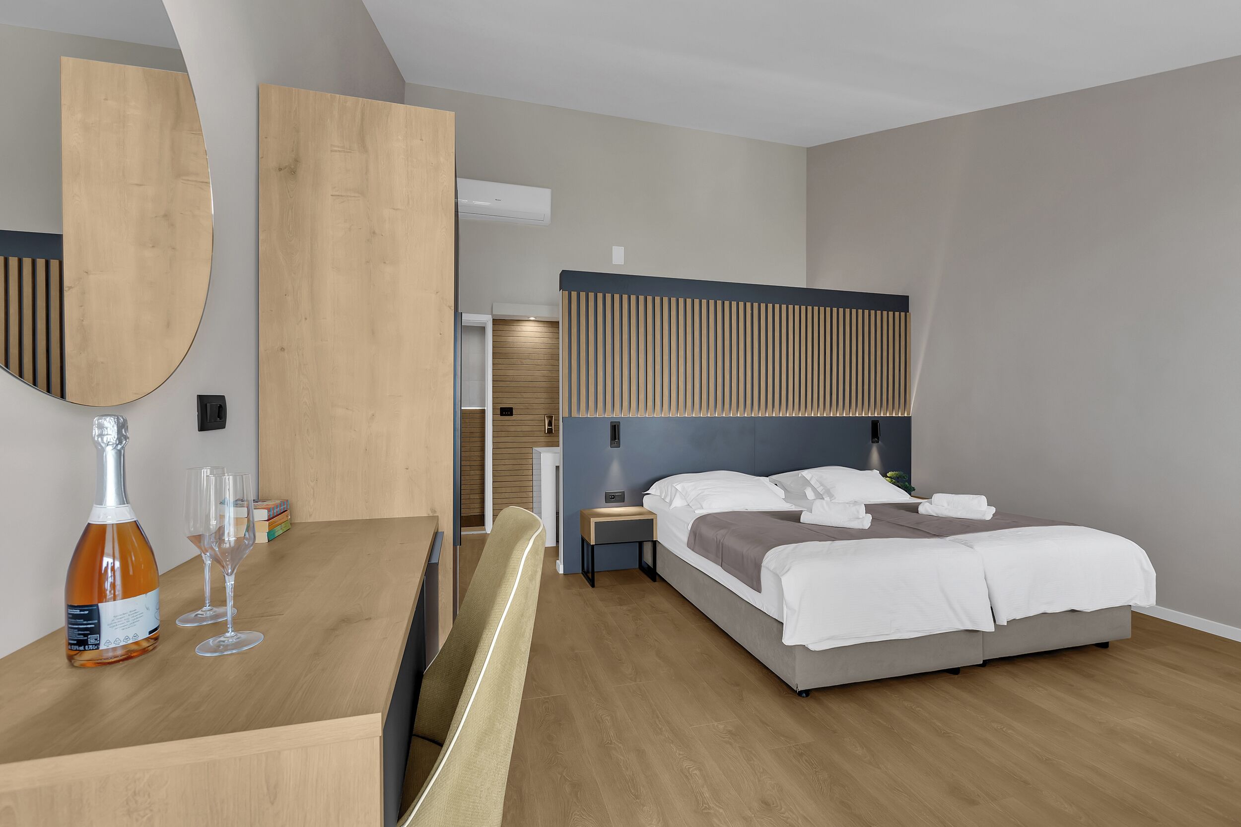 A modern hotel room with a wooden desk, a large mirror, a bed with white linens, and a nightstand with a lamp.