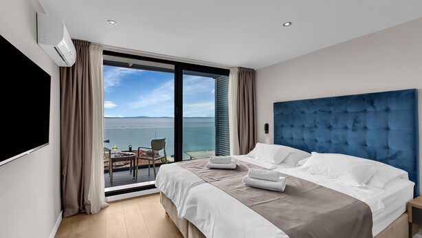 Une chambre d’hôtel moderne avec un grand lit, une tête de lit bleue et un balcon offrant une vue sur la mer.