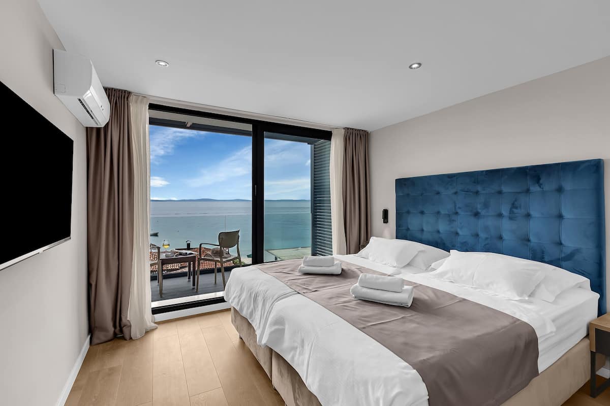 A modern hotel room with a large bed, a blue headboard, and a balcony with a view of the sea.