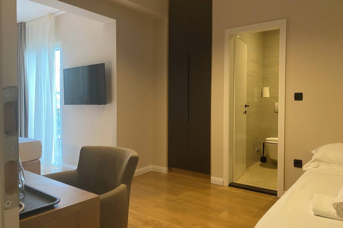 A hotel room with a wooden floor, a bed with white linens, a desk with a chair, a TV mounted on the wall, and a door leading to a bathroom.