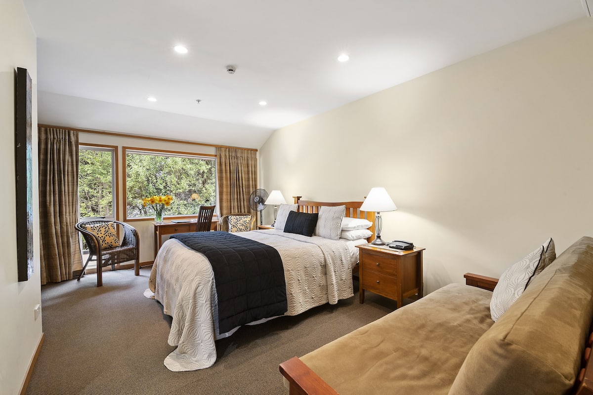 Habitación de hotel con una cama grande, mesitas de noche con lámparas, una silla y vistas a árboles por la ventana.