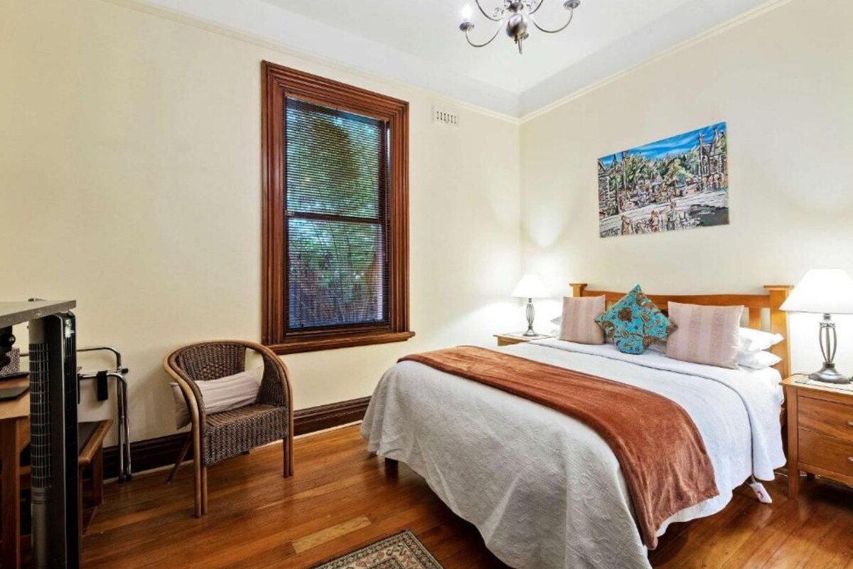 A bedroom with a large bed, a window with blinds, a chair, a bedside table with a lamp, and a painting on the wall.