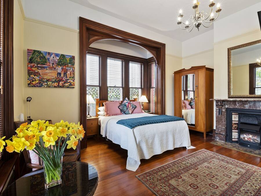 A bedroom with a large bed, a fireplace, a painting, and a window with blinds.