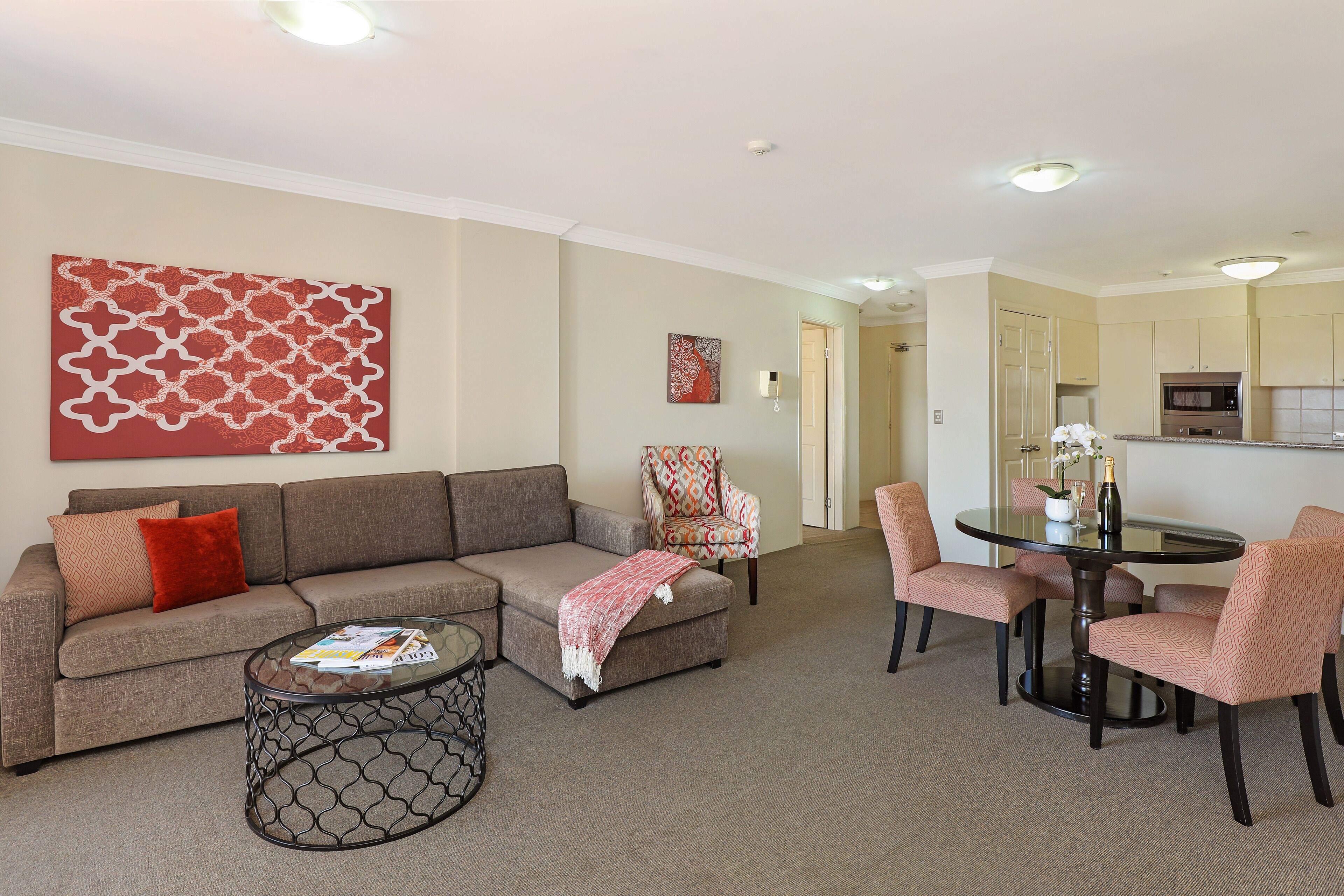 A living room with a sofa, coffee table, and patterned chairs. There is a dining table with chairs and a kitchen area in the background.