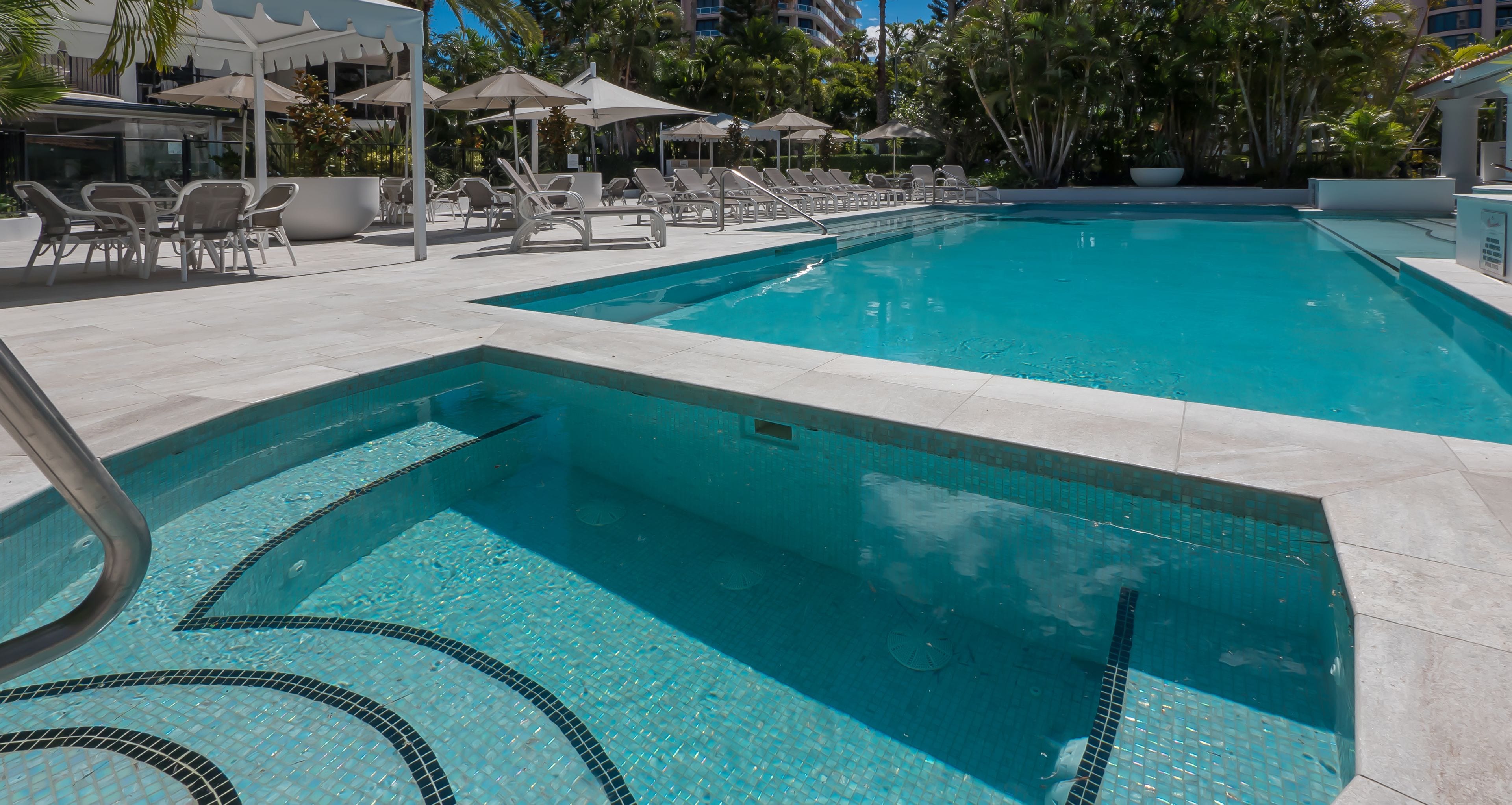 Ein Swimmingpool mit Liegestühlen und Sonnenschirmen, umgeben von Palmen und einem Gebäude im Hintergrund.