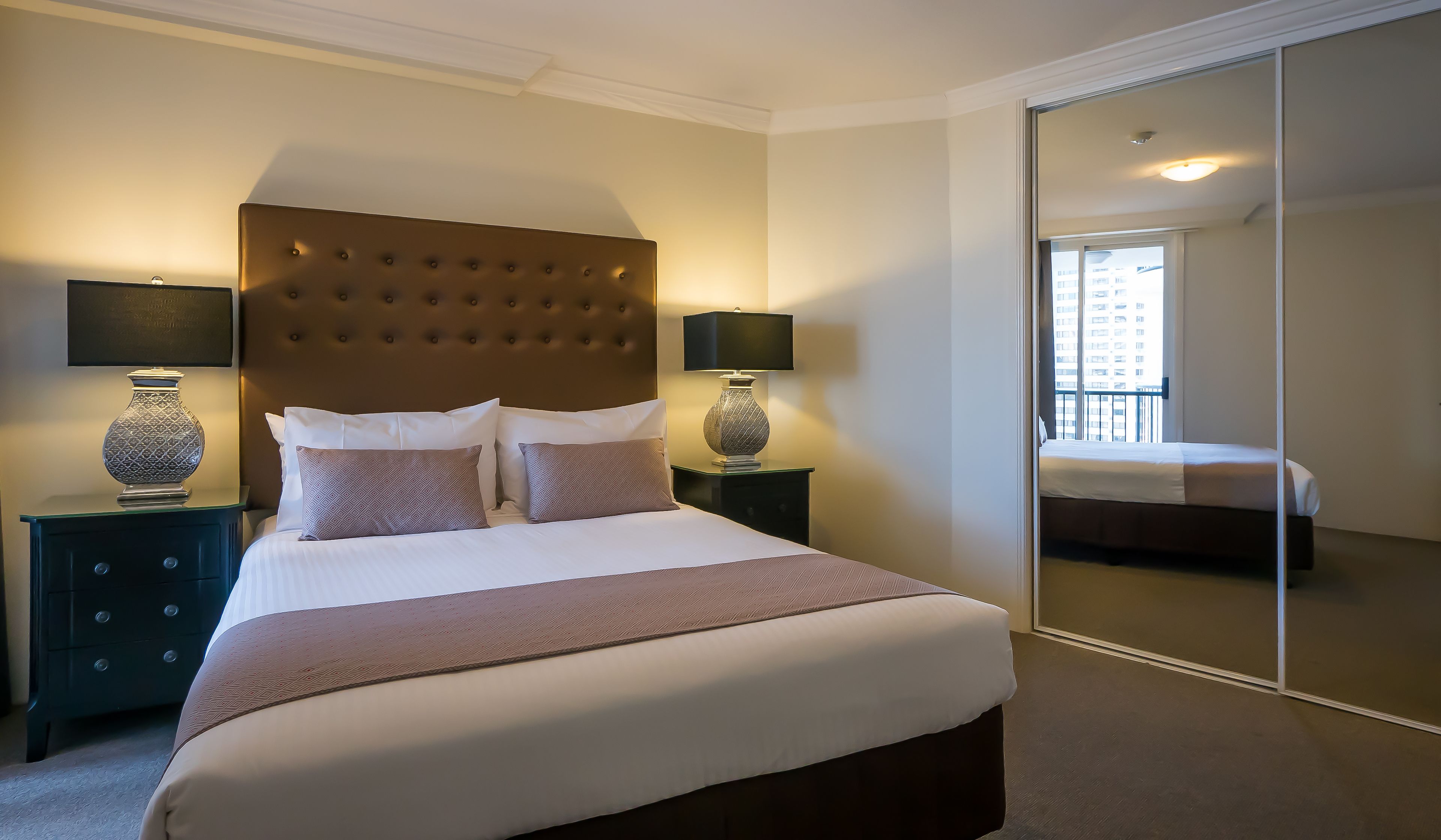 Une chambre d’hôtel avec un grand lit, deux tables de chevet avec des lampes, un grand miroir et une vue sur un immeuble par la fenêtre.