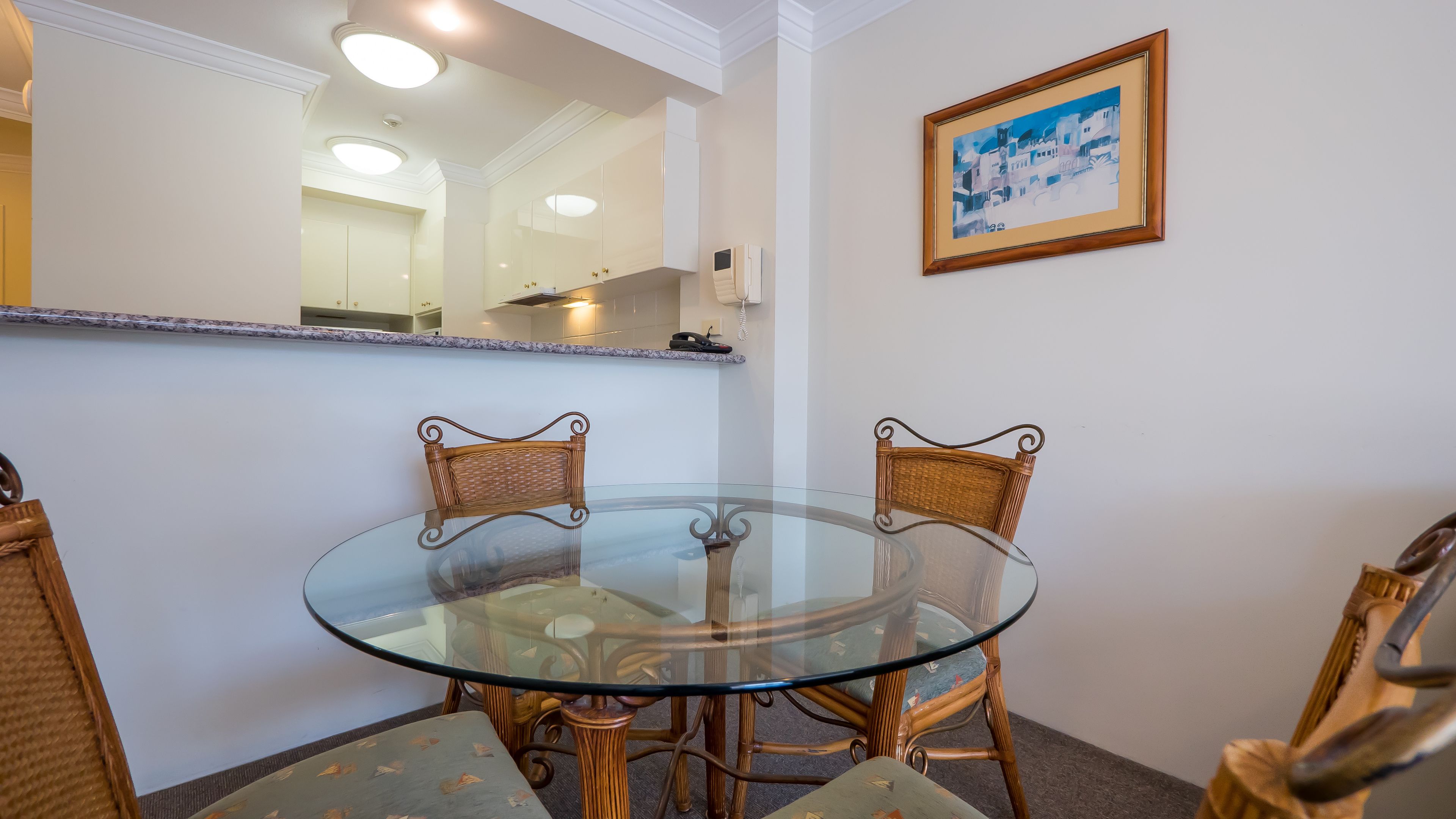 Un coin repas avec une table en verre et des chaises en osier, un tableau encadré au mur et une cuisine en arrière-plan.