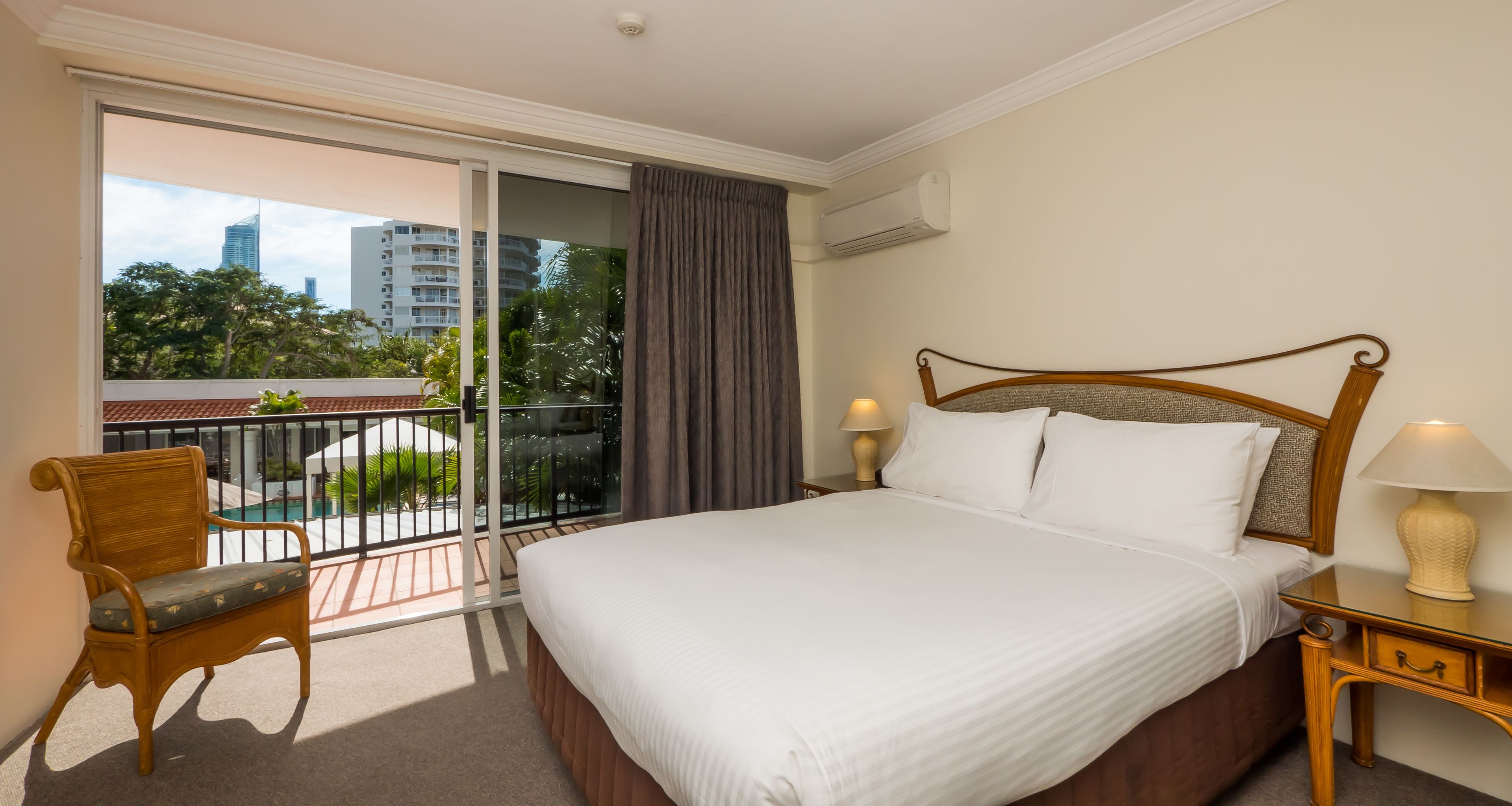 Une chambre d’hôtel avec un grand lit, une chaise, une table de chevet et une vue sur un balcon donnant sur une piscine et des immeubles.
