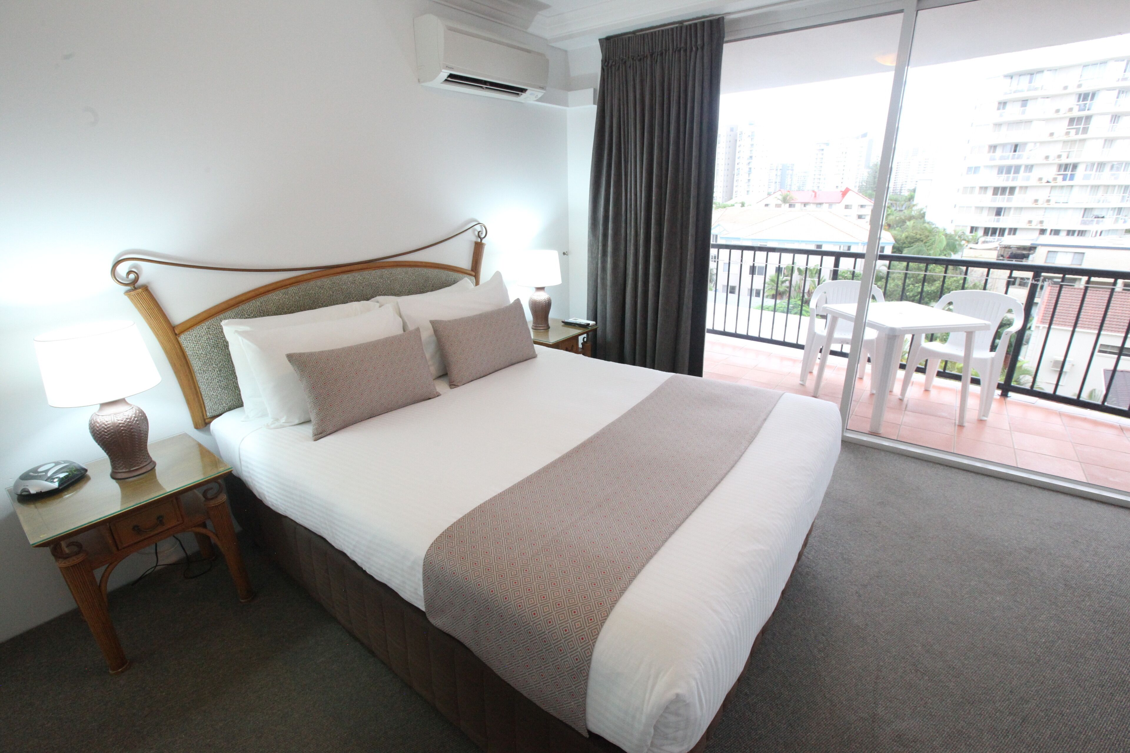 Habitación de hotel con una cama grande, una mesita de noche con lámpara, una ventana con vistas a edificios y un balcón con sillas.