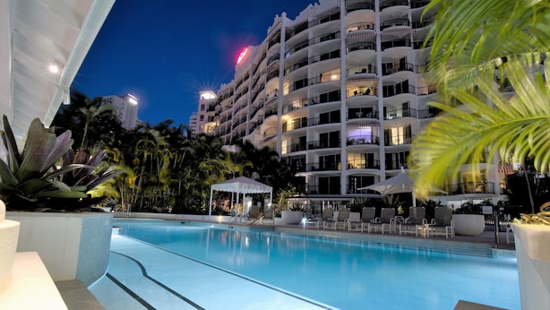 A swimming pool with lounge chairs and umbrellas, surrounded by palm trees and a multi-story building with balconies. - Marrakesh Apartments (Surfers Paradise)