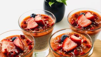 Vier Gläser mit mehrschichtigen Desserts, belegt mit Erdbeeren und Blaubeeren, auf einer Holzoberfläche. Im Hintergrund steht ein Blumentopf.