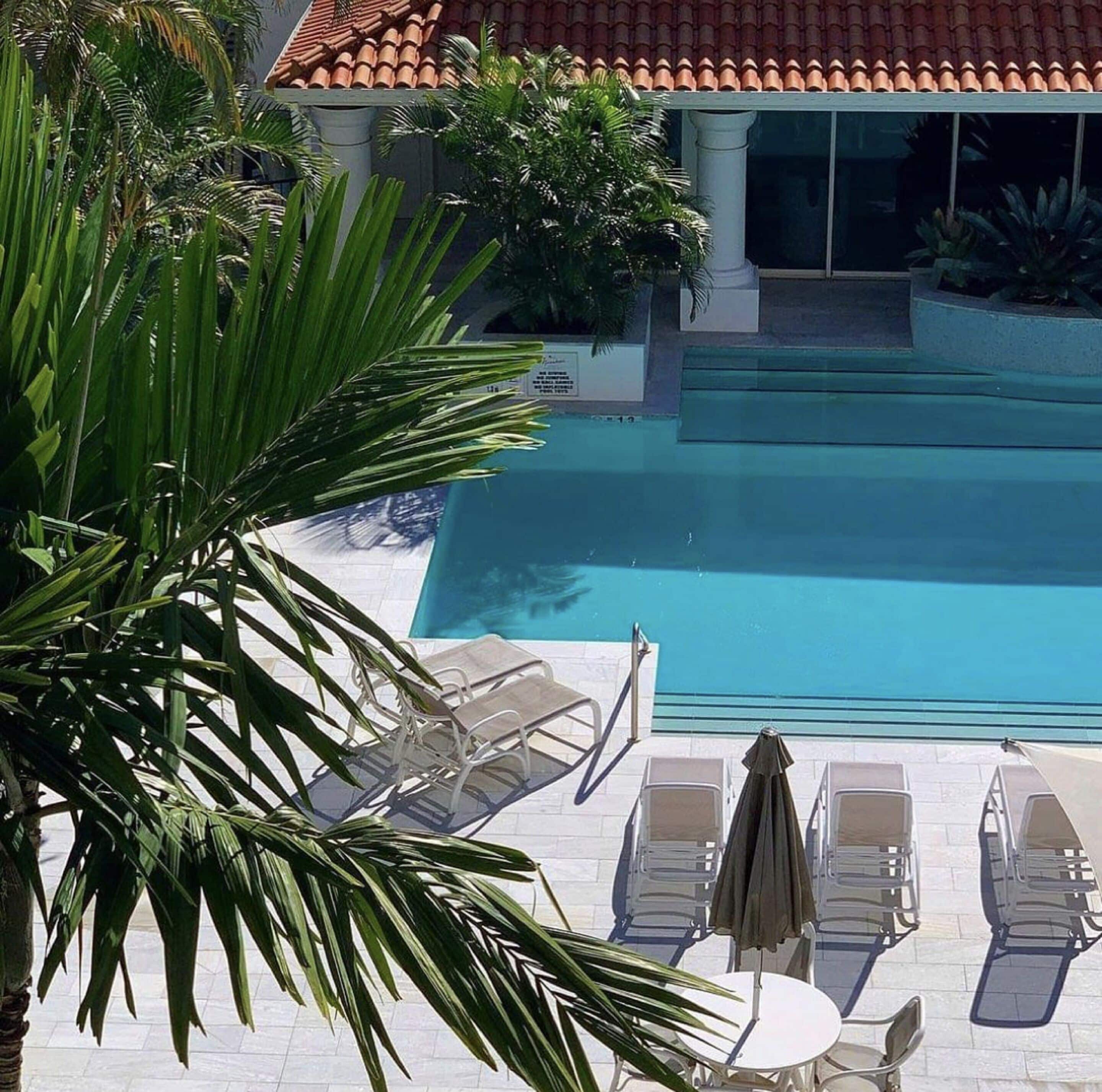 Una piscina con sillas de descanso y sombrillas, rodeada de palmeras y un techo de tejas.