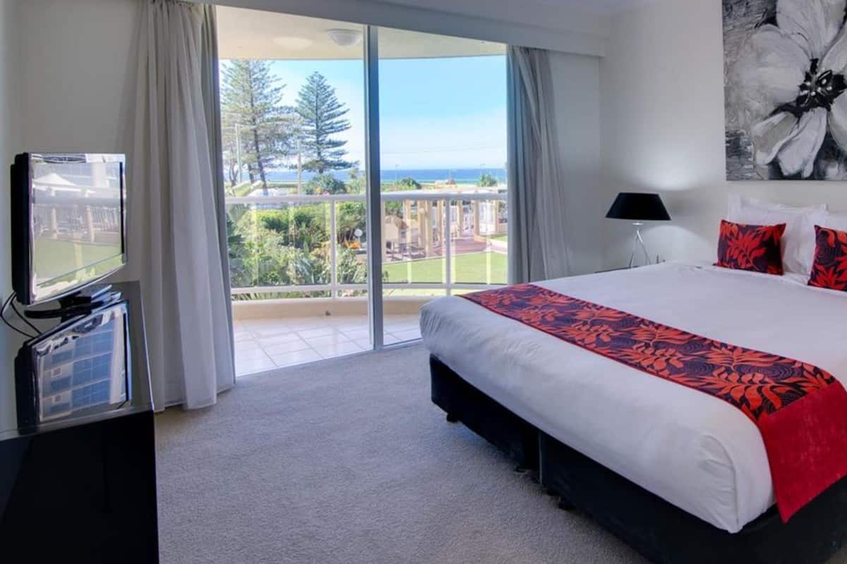 A hotel room with a large bed, a TV, a balcony with a view, and a painting on the wall.