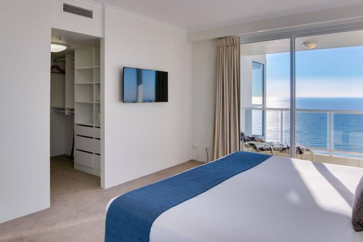 A hotel room with a bed, a TV, a closet, and a balcony with a view of the sea.