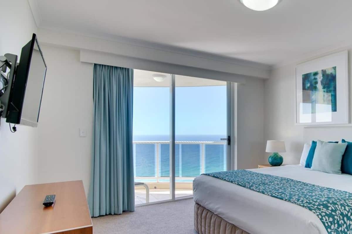 A hotel room with a bed, a desk, a TV, and a balcony with a view of the sea.