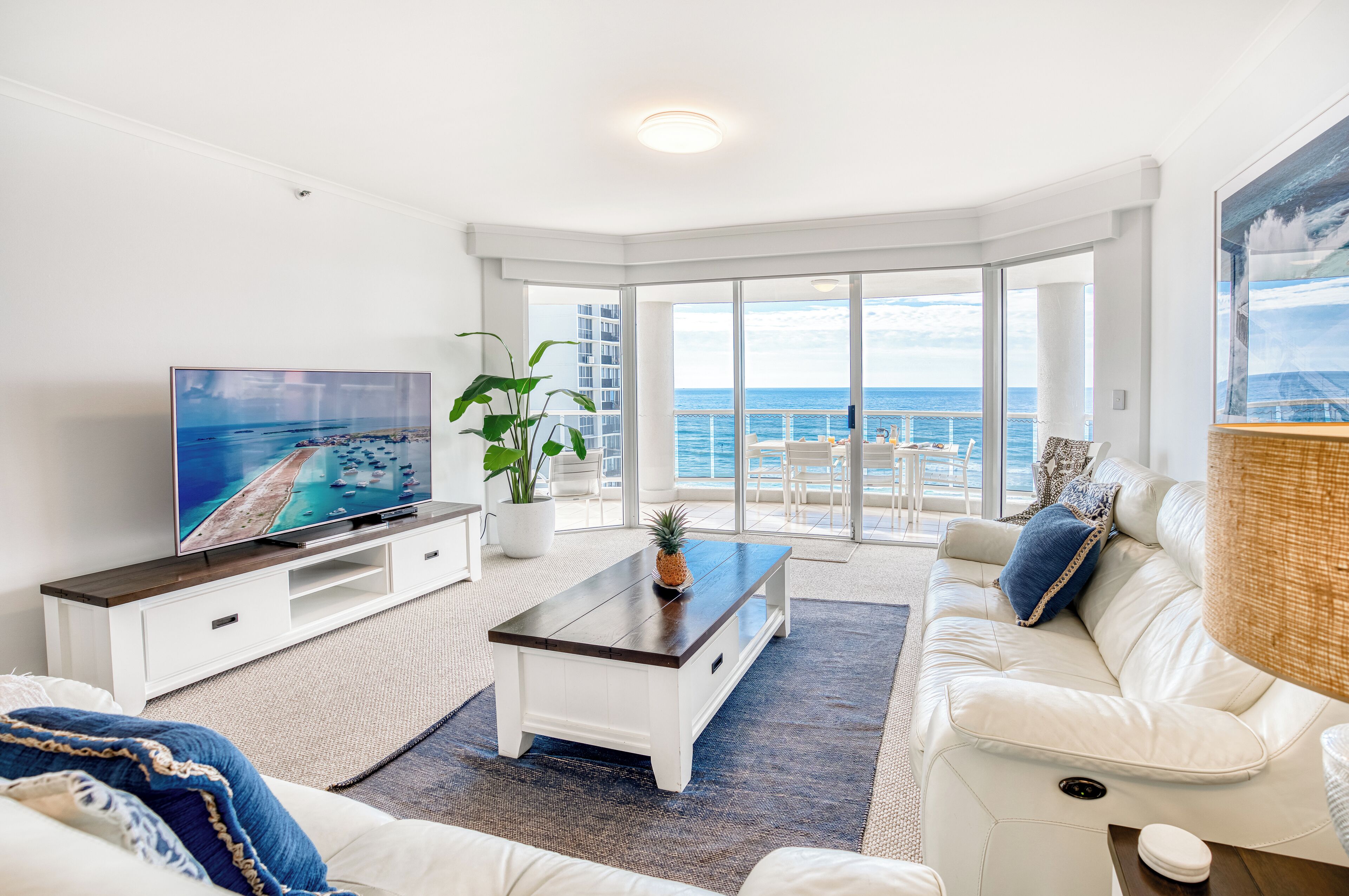 A living room with a TV, white sofa, and a large window with a view of the ocean.