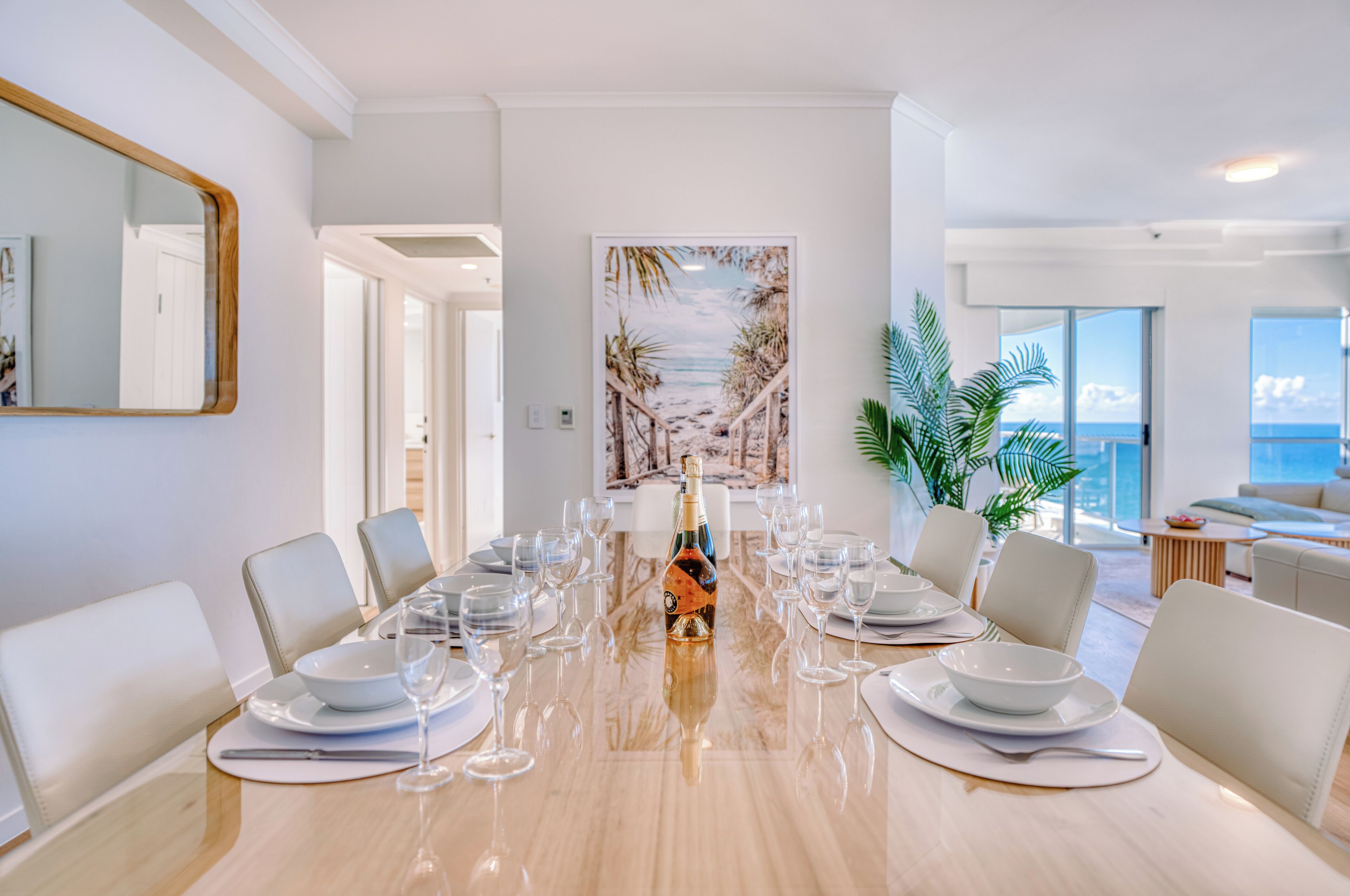 A dining area with a long table set for a meal, a bottle of champagne, and a painting of a beach scene on the wall.