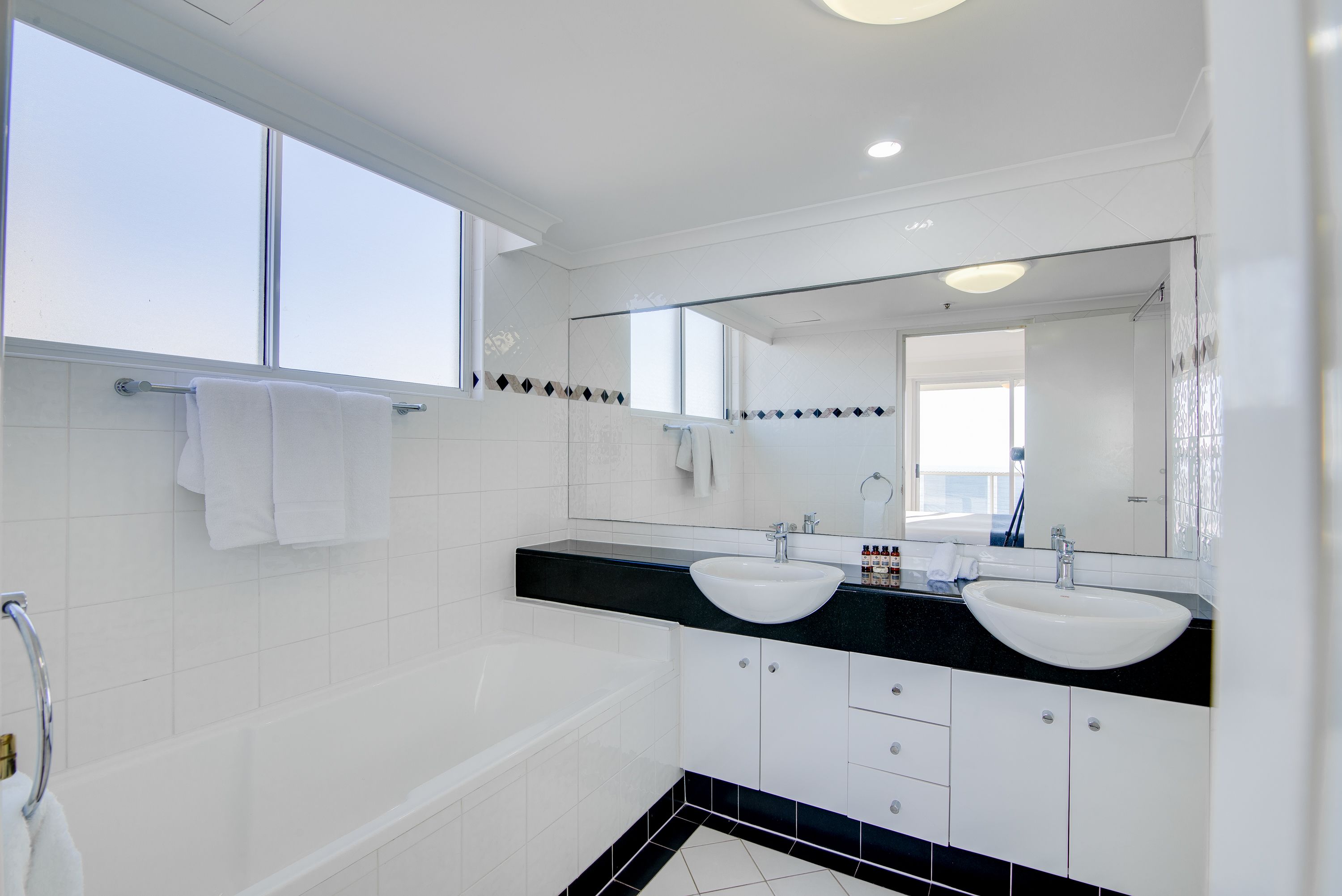 A modern bathroom with a bathtub, two sinks, a large mirror, and a window.