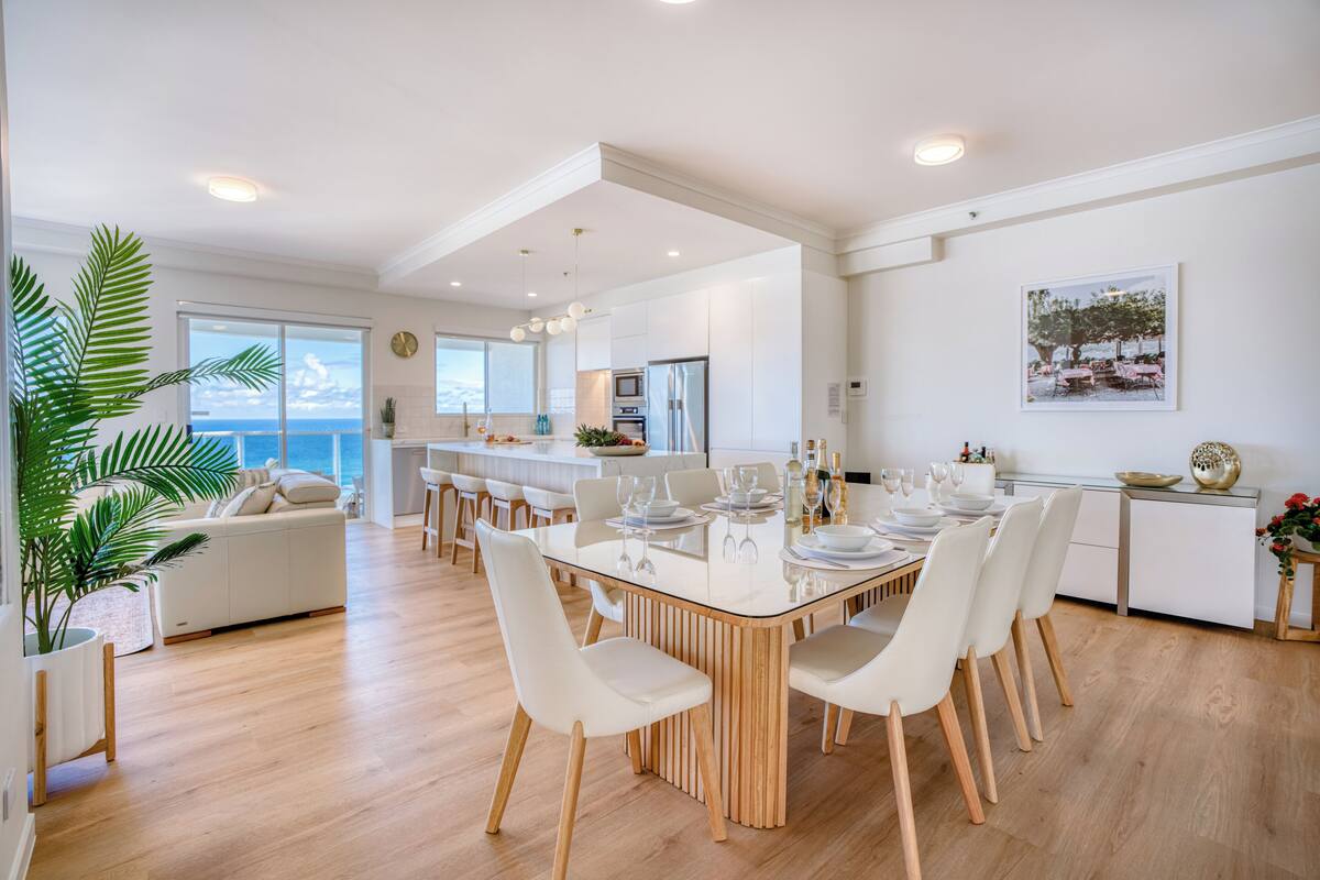 A modern kitchen with white cabinets, a dining area with a large table set for six, and a view of the ocean through large windows.