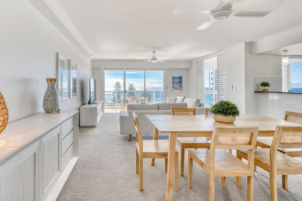 A modern living room with a dining area, large windows, and a view of the ocean.