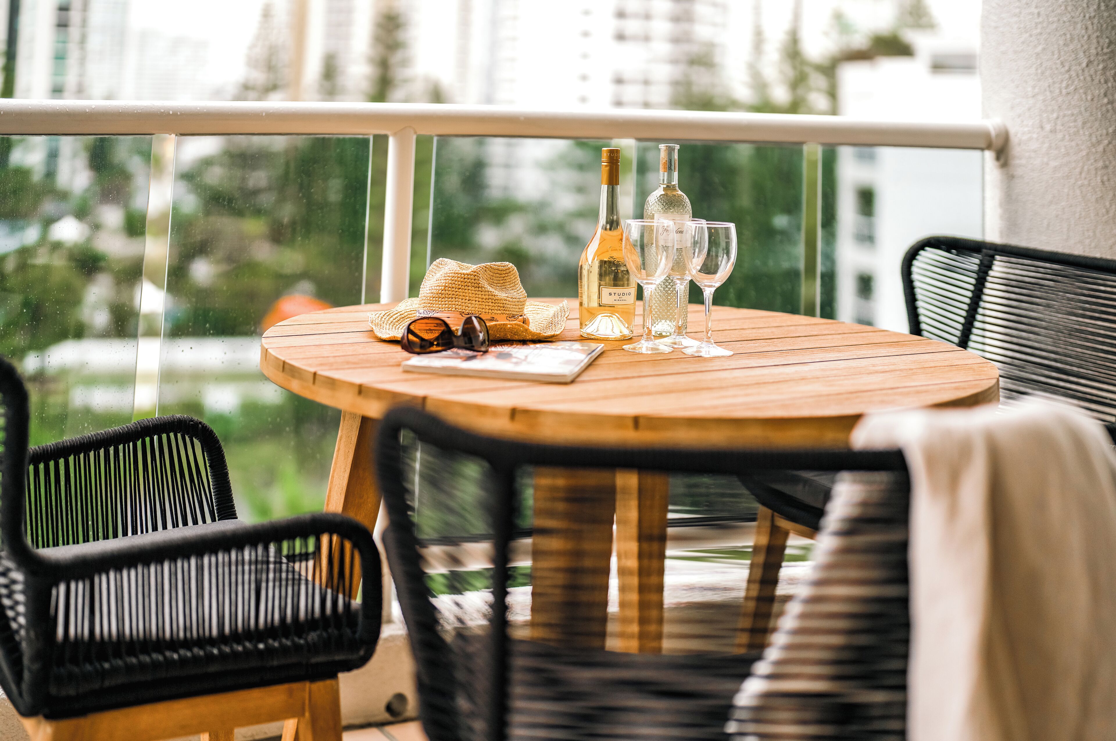 A round wooden table with a bottle of wine, two wine glasses, a straw hat, and a pair of sunglasses on a balcony with black wicker chairs.