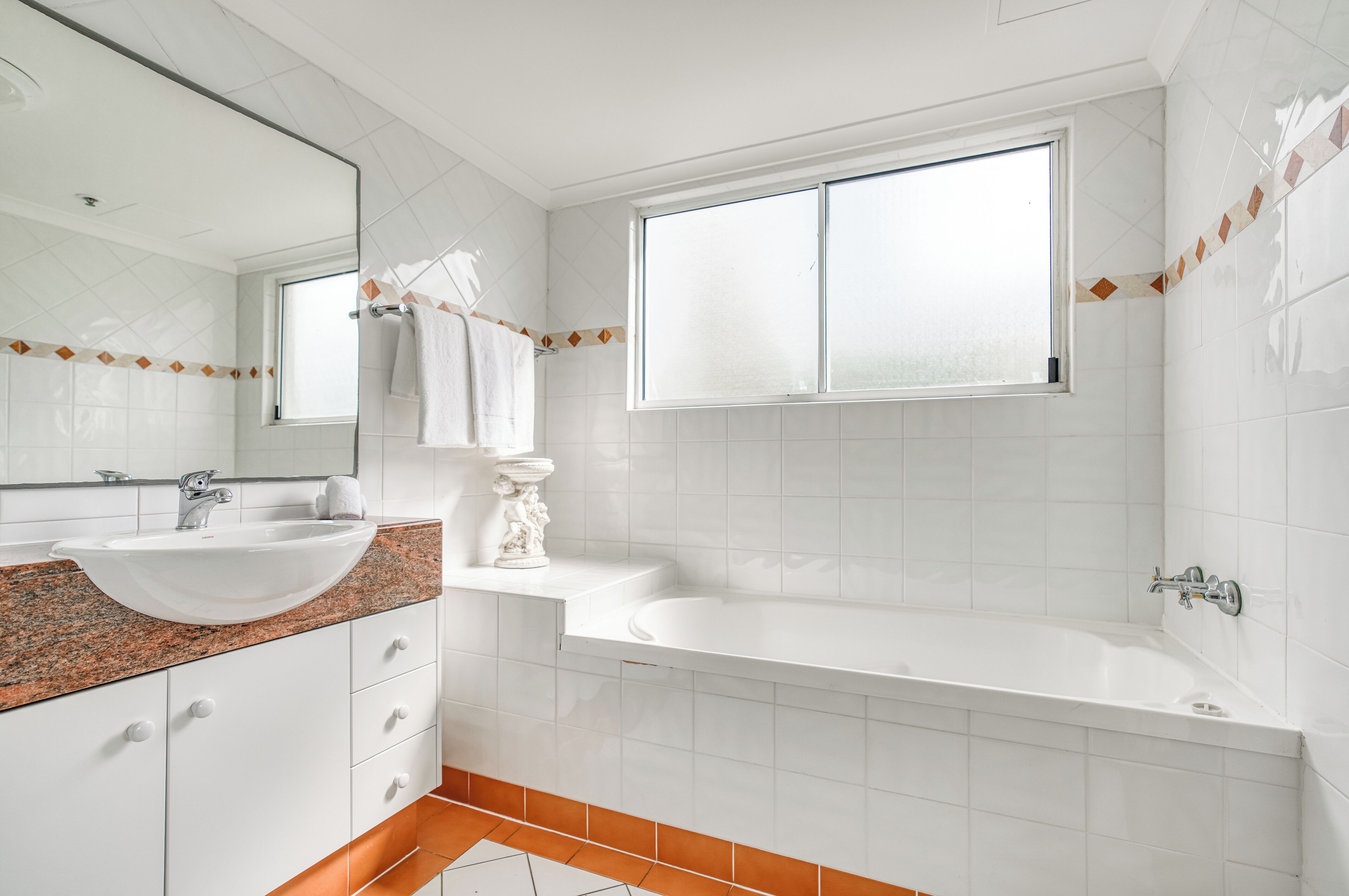 Een moderne badkamer met een wit wastafelmeubel, wastafel en bad. Er is een raam dat zorgt voor natuurlijk licht en een decoratieve tegelrand.