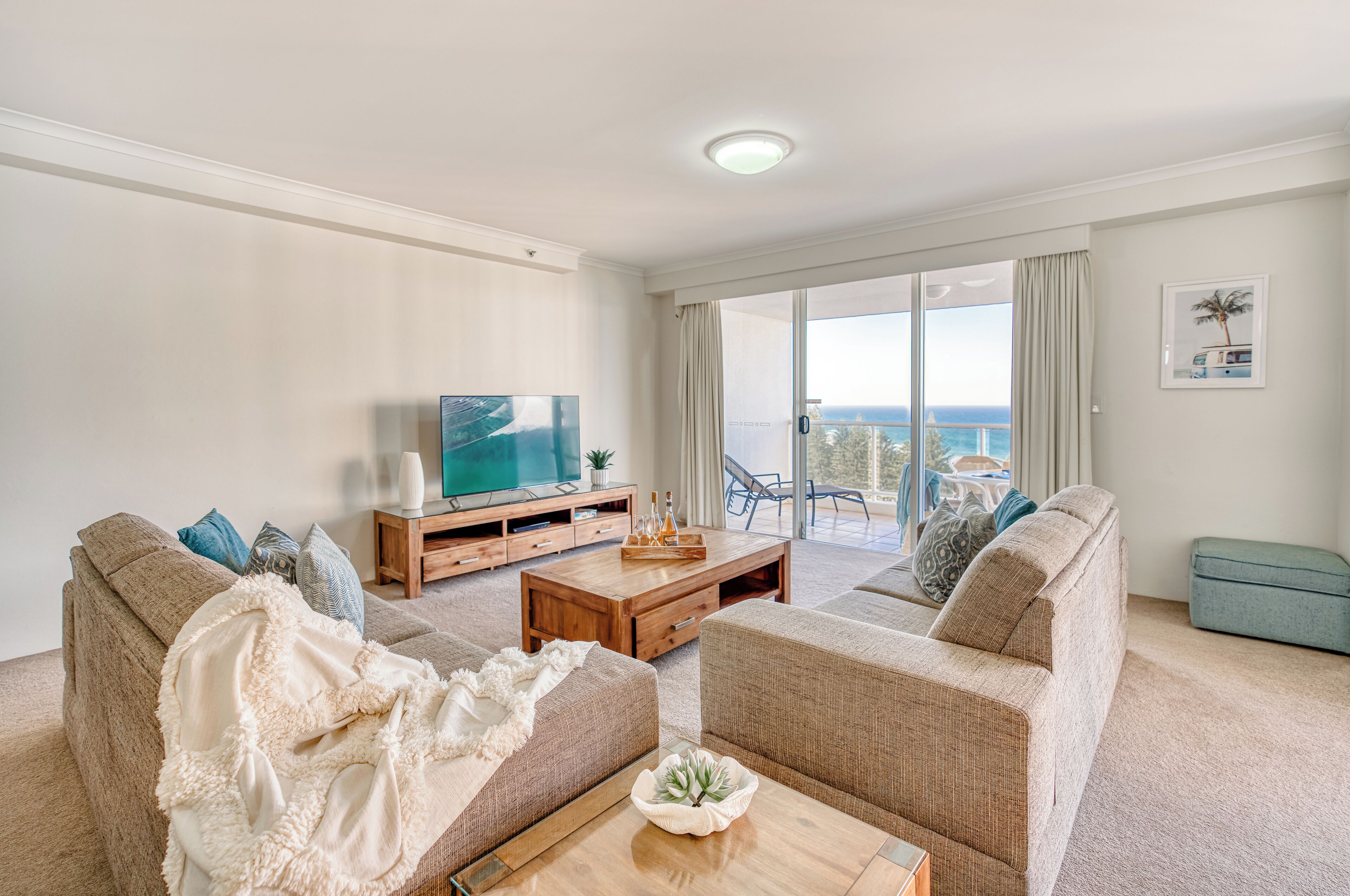 Een woonkamer met een bank, salontafel en televisie, een balkon met uitzicht op zee en een kleine tafel met flessen.