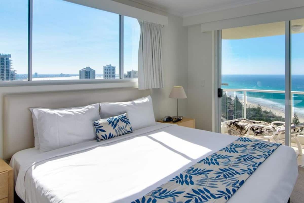 A hotel room with a large bed, a nightstand, a lamp, a TV, and a balcony with a view of the ocean and buildings.