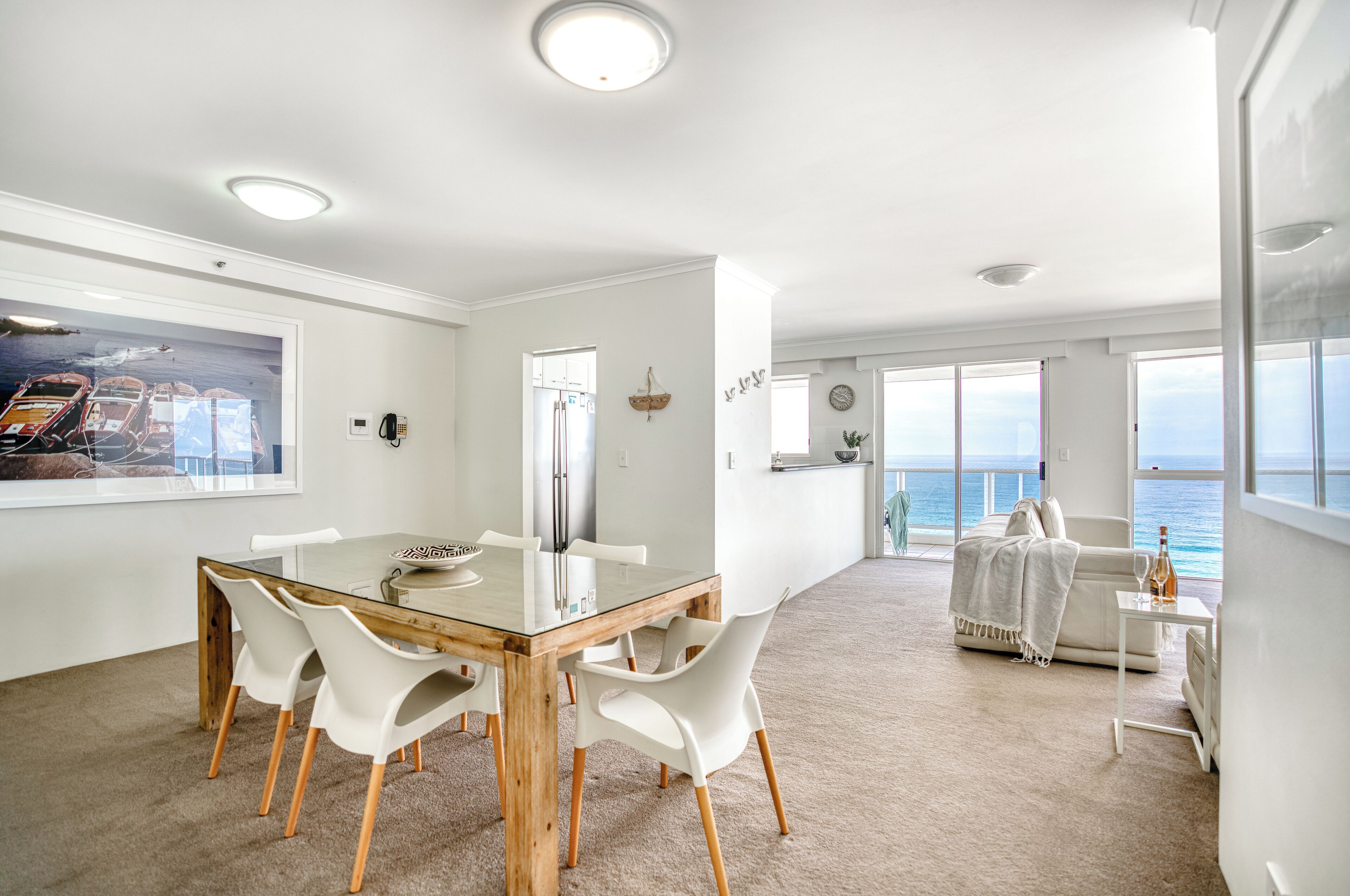 A spacious room with a large window overlooking the sea, a dining area with a glass table and white chairs, and a cozy seating area with a view.