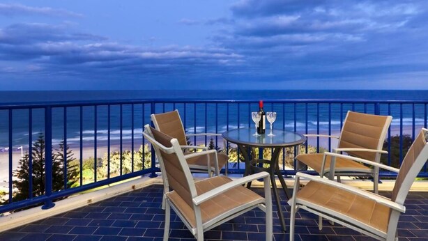 A balcony with a view of the ocean, a round table with chairs, and a bottle of wine.