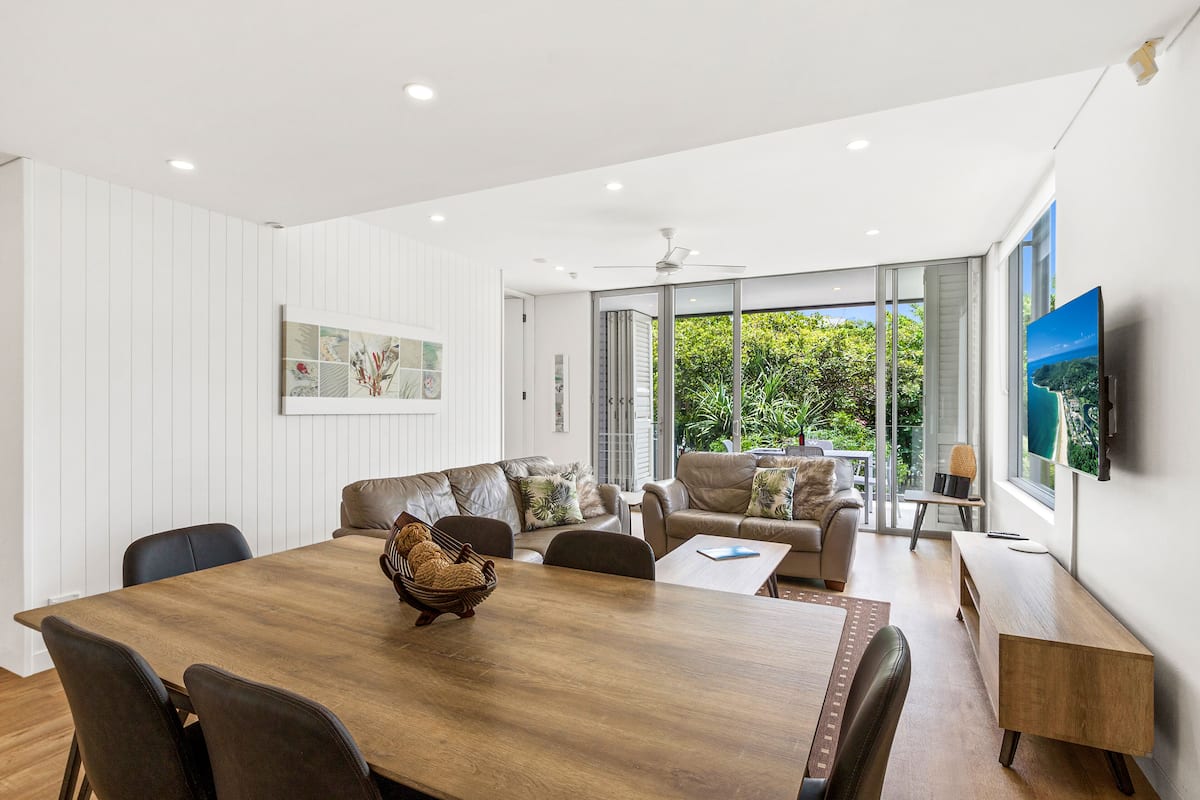 A modern living room with a wooden dining table, leather sofas, and a large TV.