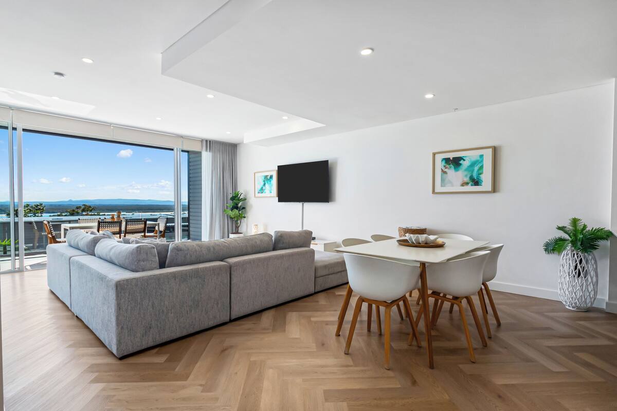 A modern living room with a grey sectional sofa, a dining table with white chairs, a flat-screen TV mounted on the wall, and a large window offering a view of the outdoors.