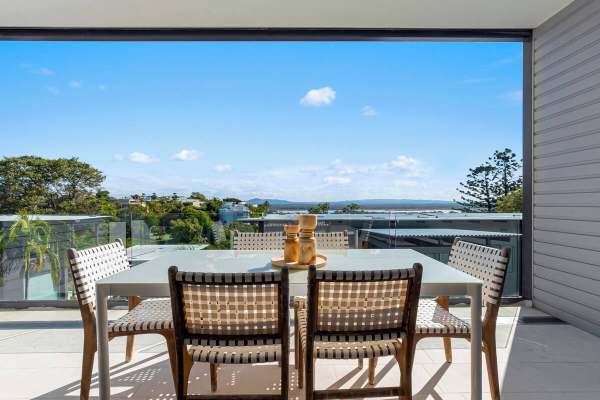 A dining area with a glass table and wicker chairs, overlooking a cityscape with buildings and greenery.