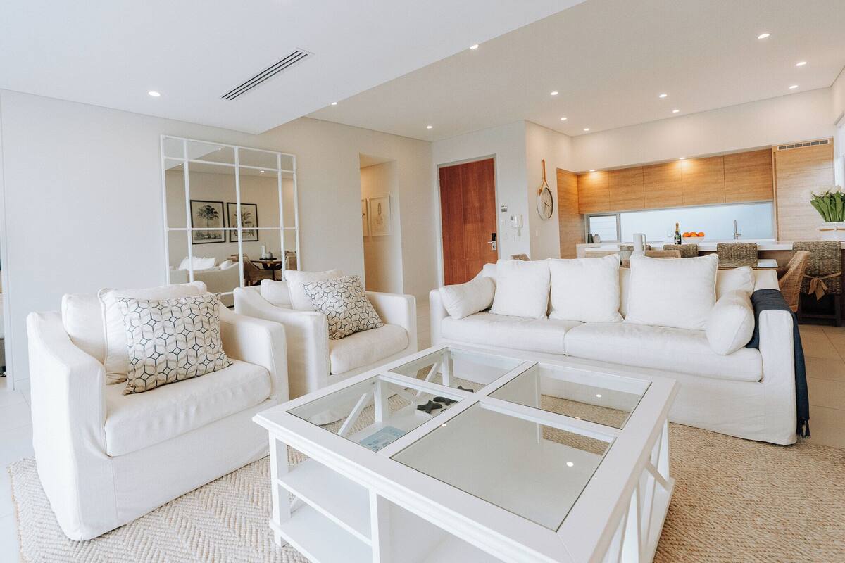 A modern living room with white furniture, a glass coffee table, and a kitchen area in the background.