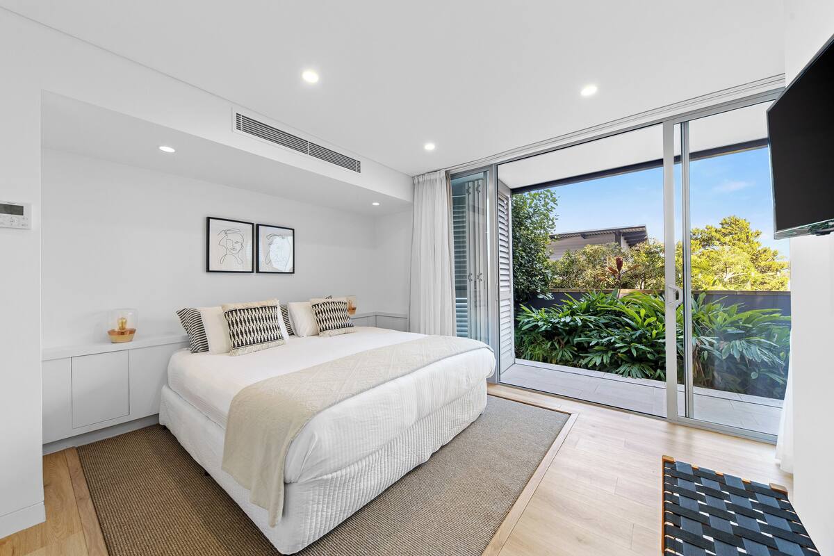 A modern bedroom with a large bed, a sliding glass door to a balcony, and a view of greenery outside.
